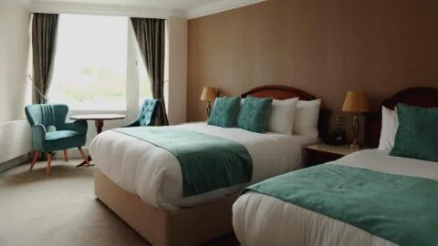 Hotel room with two double beds, teal accents, a small round table with two chairs by the window, bedside lamps, and neutral-toned walls.