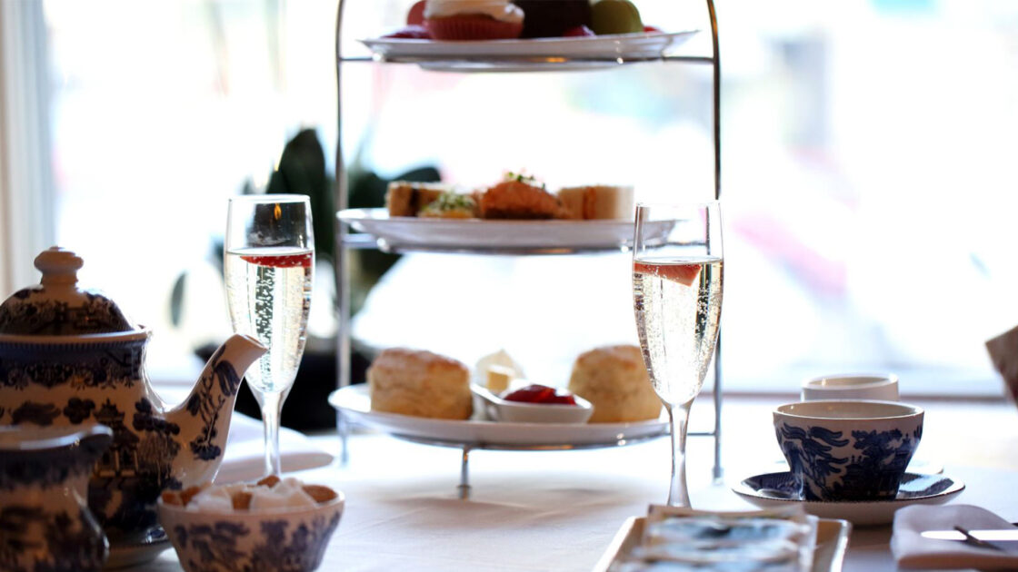 A table set for afternoon tea with a teapot, cups, two glasses of sparkling wine, and a tiered tray holding pastries, sandwiches, and scones.