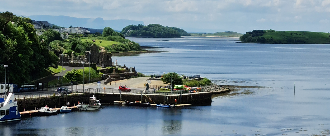 Small boats docked at a harbor with a parking area, grassy ruins, and houses nearby, overlooking a wide river or bay with distant green hills under a partly cloudy sky.