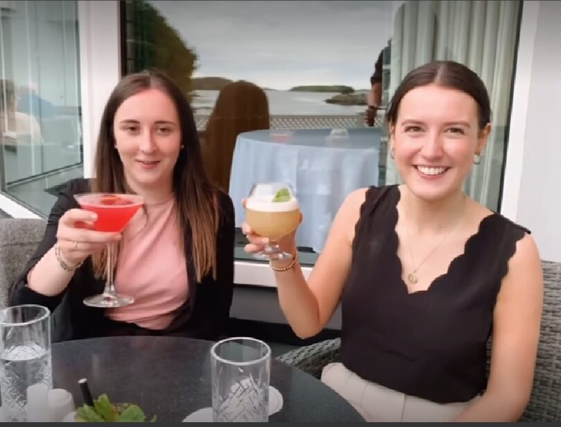 Two women are sitting at an outdoor table, holding up cocktails and smiling at the camera, with a scenic view in the background.