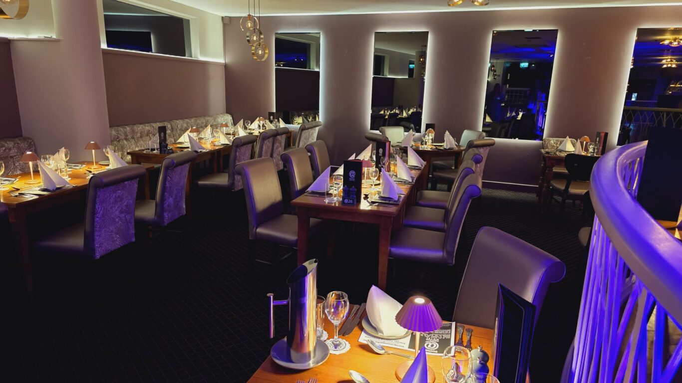 A modern, dimly lit restaurant dining area with neatly set tables, upholstered chairs, menus, glasses, and napkins arranged for guests.