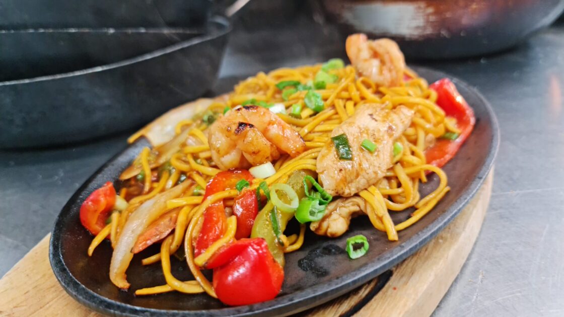 A plate of stir-fried noodles with shrimp, chicken, bell peppers, onions, and green onions, served on a sizzling black dish.