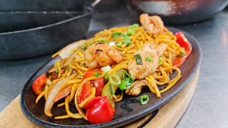 A plate of stir-fried noodles with shrimp, chicken, bell peppers, onions, and green onions, served on a sizzling black dish.