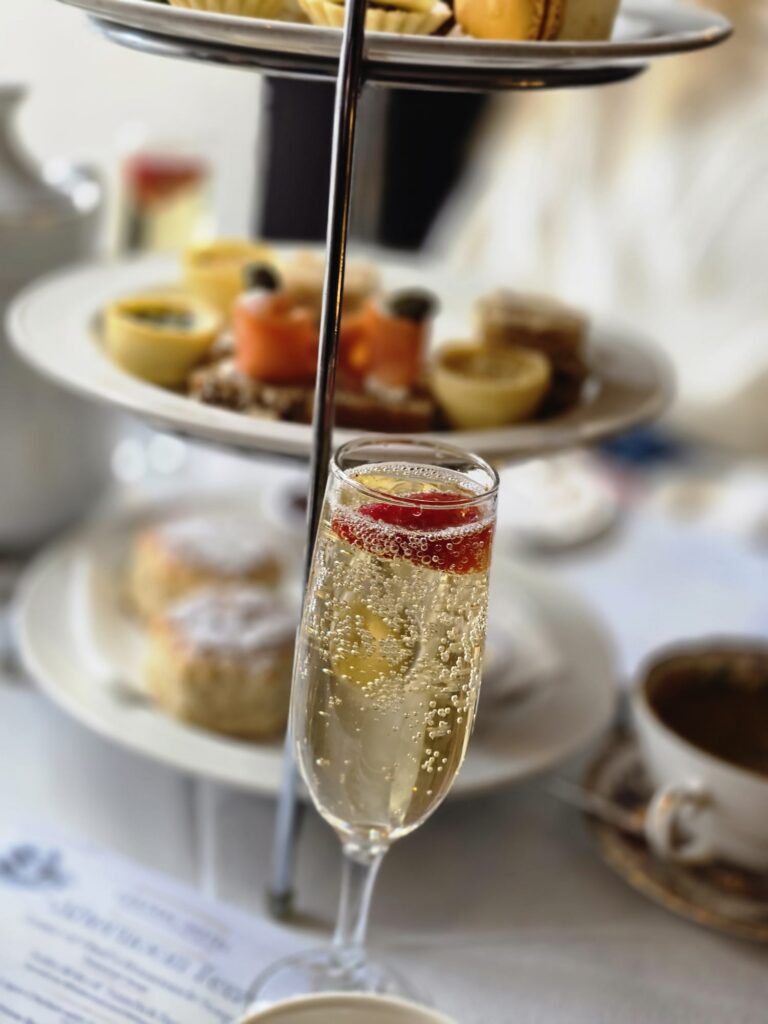 WhatsApp Image 2026-02-27 at 15.59.46 A glass of sparkling wine with a berry garnish is in focus, set in front of a tiered tray of assorted afternoon tea pastries and sandwiches.