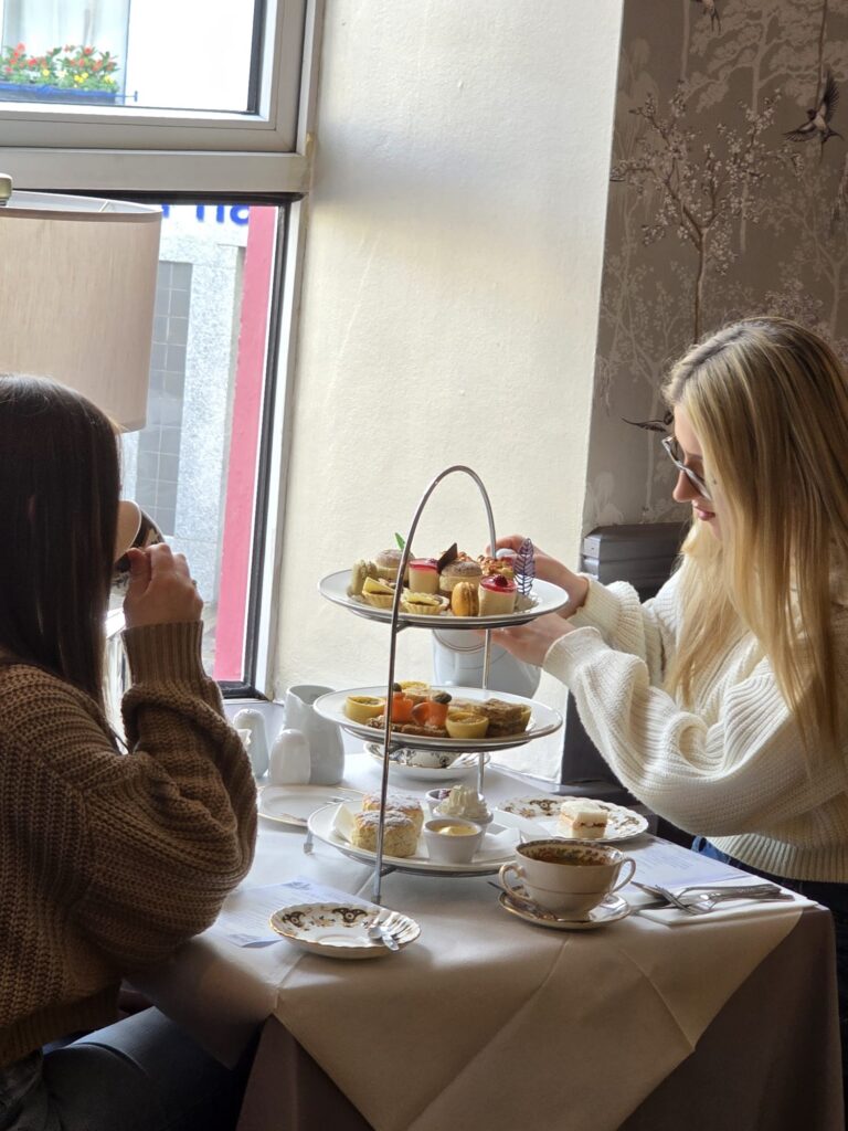 WhatsApp Image 2026-02-27 at 15.59.49 (2) Two women sit at a table by a window, enjoying afternoon tea with a three-tiered tray of pastries and cups of tea.
