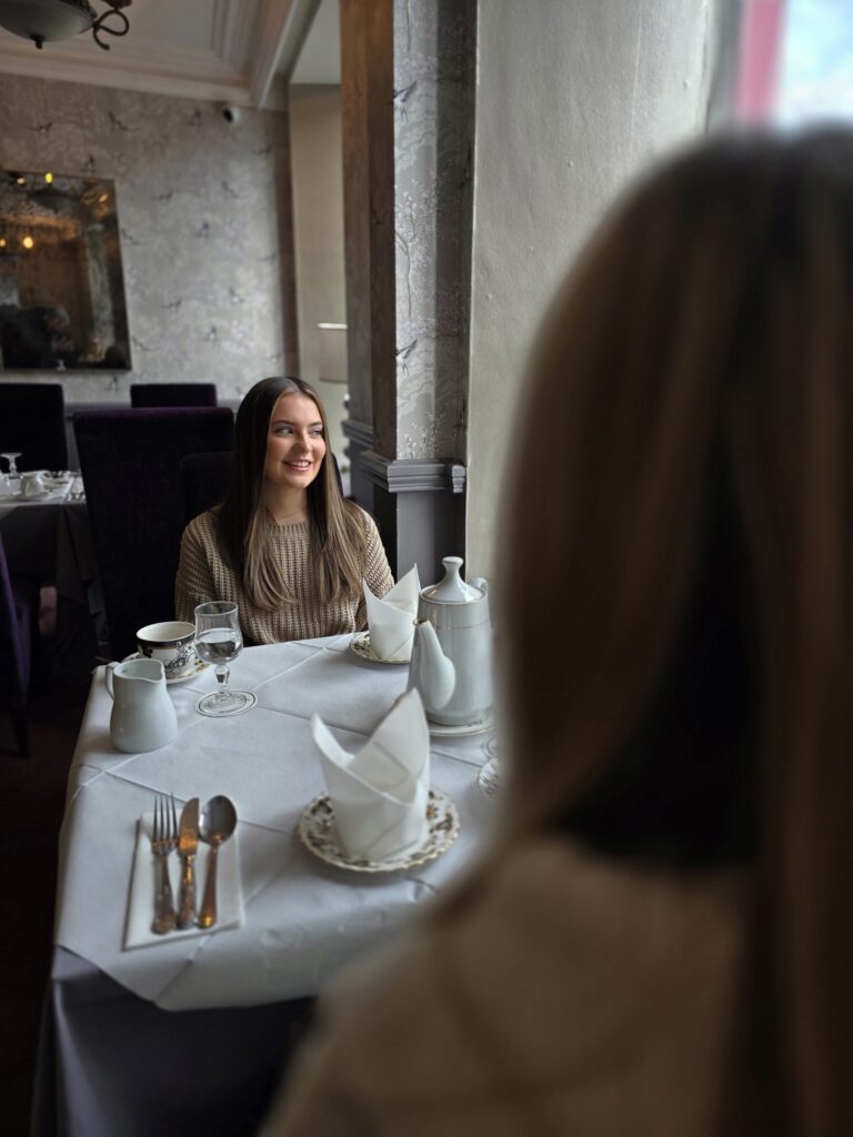 WhatsApp Image 2026-02-27 at 15.59.49 (8) A woman sits at a restaurant table set for two, smiling at another person whose back is to the camera. Tableware and folded napkins are arranged neatly.