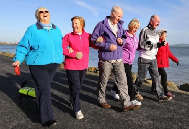 Six older adults walk arm-in-arm along a waterfront path on a sunny day, smiling and laughing together.