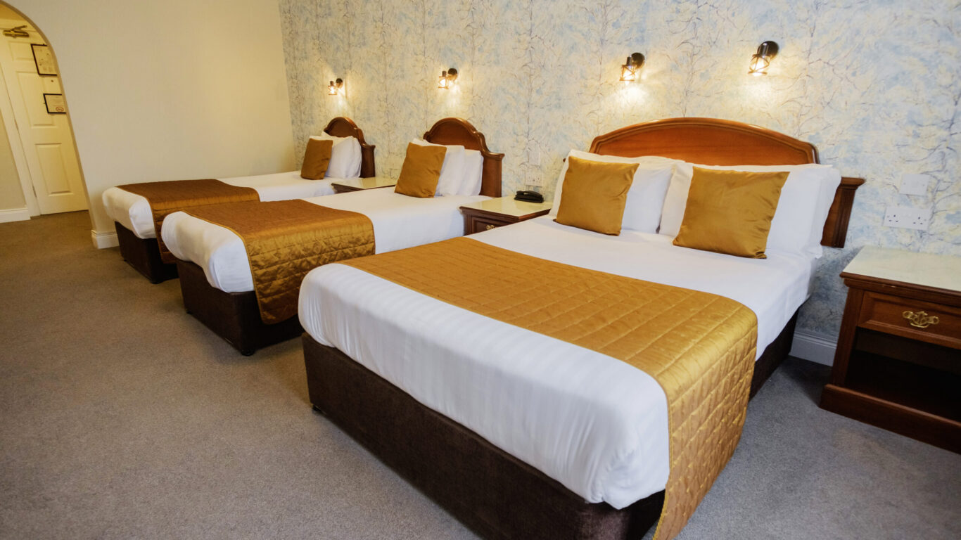 Hotel room with two double beds and one single bed, each with white linens and gold bed runners, against a patterned wall with two wall-mounted lights.