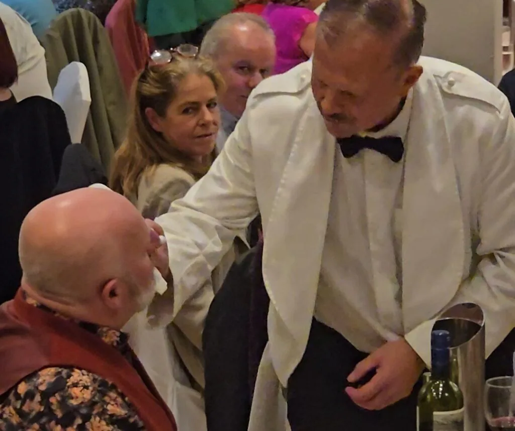 WhatsApp Image 2026-05-13 at 16.03.08 (1) A waiter in a white jacket and bow tie gestures toward a seated, bald man at a formal dining event; others are seated around them and wine is on the table.