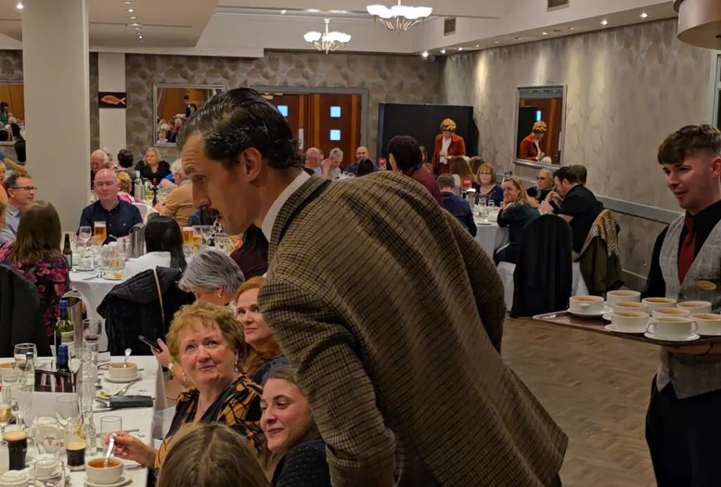 WhatsApp Image 2026-05-13 at 16.03.08 A man in a brown plaid jacket leans over to speak with seated guests at a crowded formal dinner event; a waiter carries a tray of soup bowls nearby.