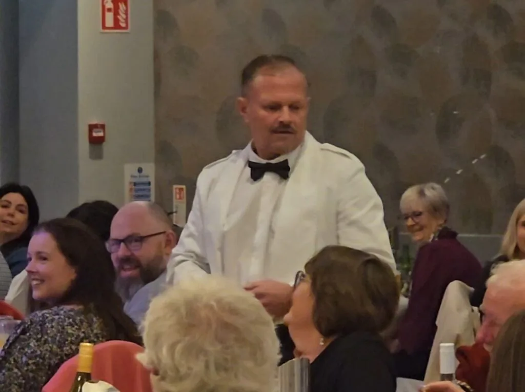 WhatsApp Image 2026-05-13 at 16.03.08 (2) A man in a white jacket and black bow tie stands among seated people at a formal event; some attendees are smiling and talking.